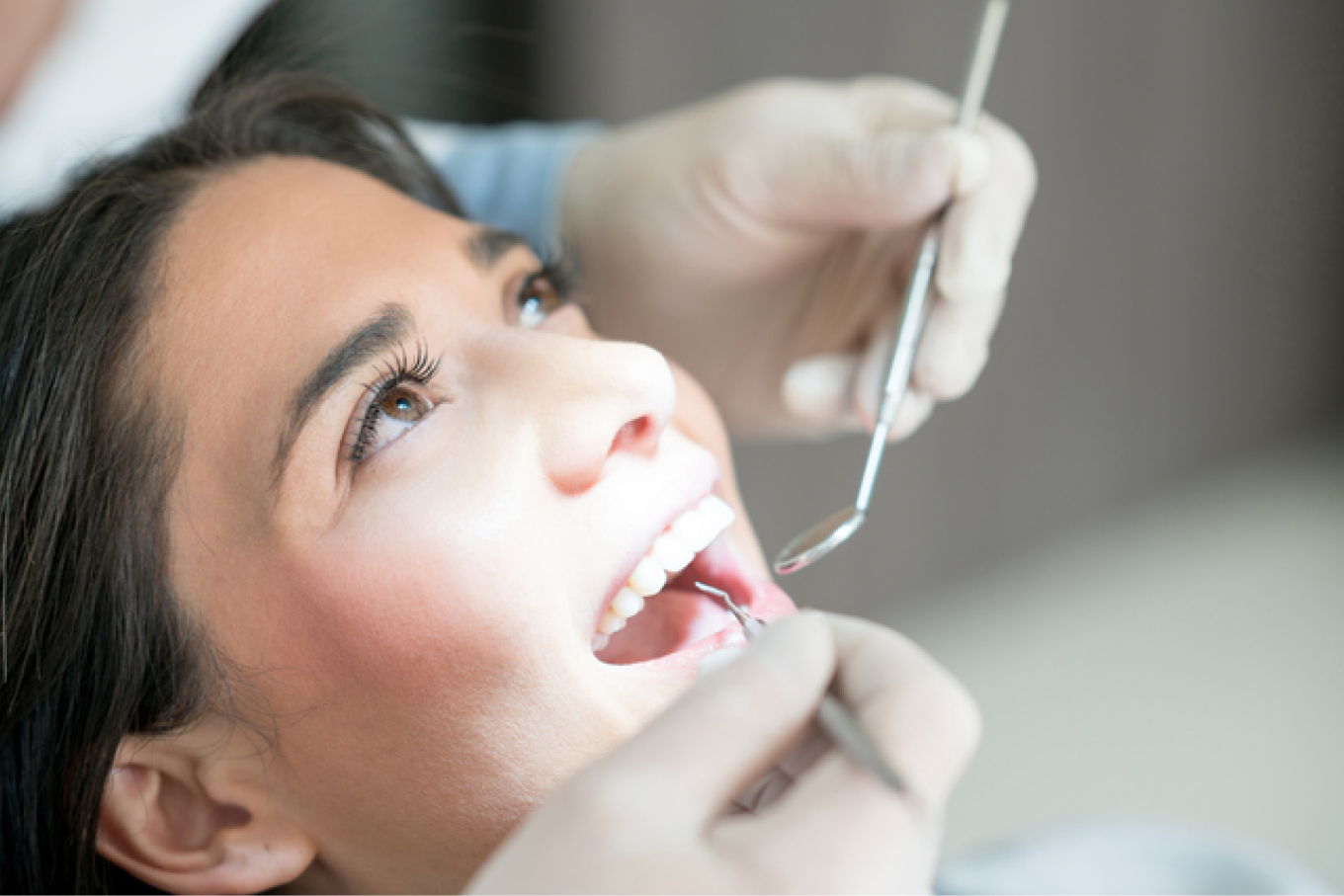 Close-up of a woman receiving a dental examination with a dentist's gloved hands holding dental tools near her open mouth.