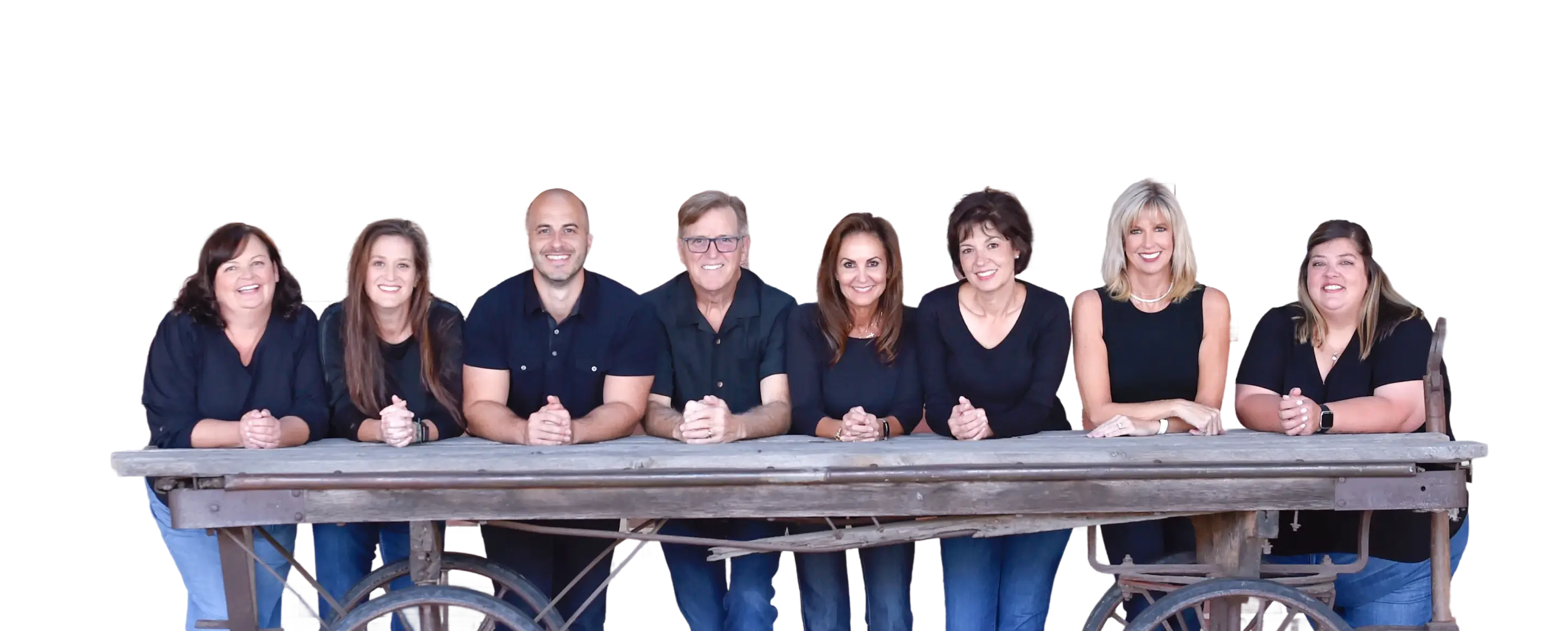 Eight smiling adults leaning on a rustic wooden table, all wearing dark tops and blue jeans.