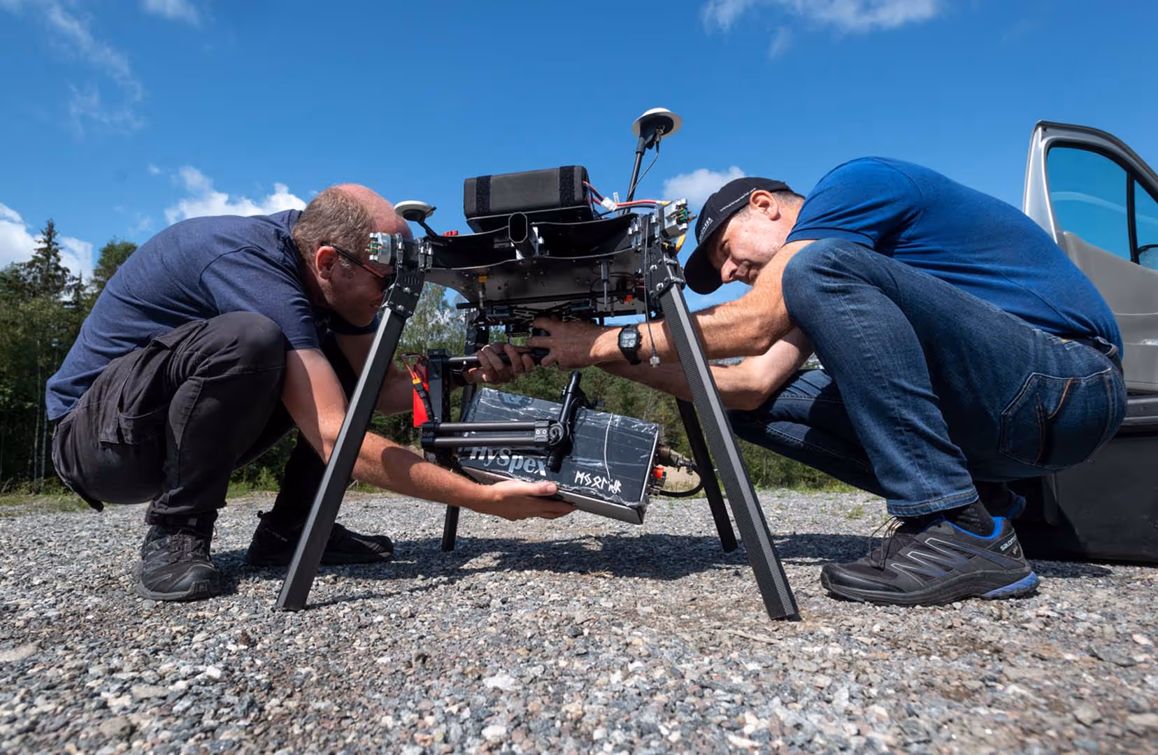 Two men crouched on gravel working on installing equipment inside a black four-legged drone under a blue sky.