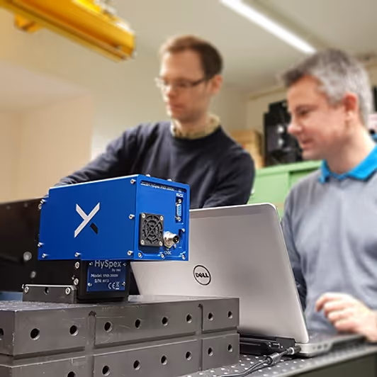 Two men working with a blue HySpex VNIR 1600 hyperspectral imaging device next to a Dell laptop in a lab setting.