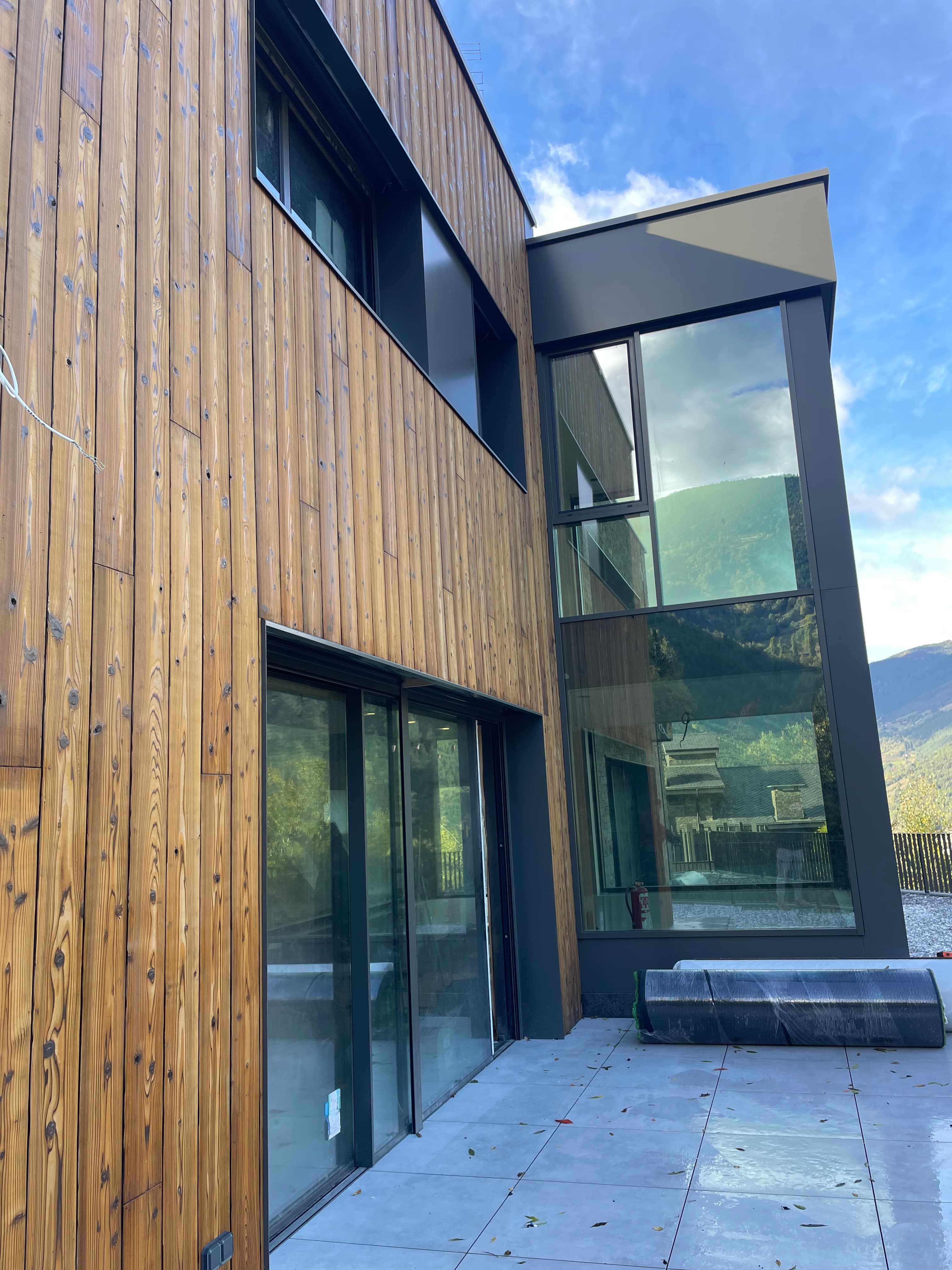 Façana moderna amb revestiment de fusta i grans finestres de vidre que reflecteixen un paisatge muntanyós sota un cel blau.