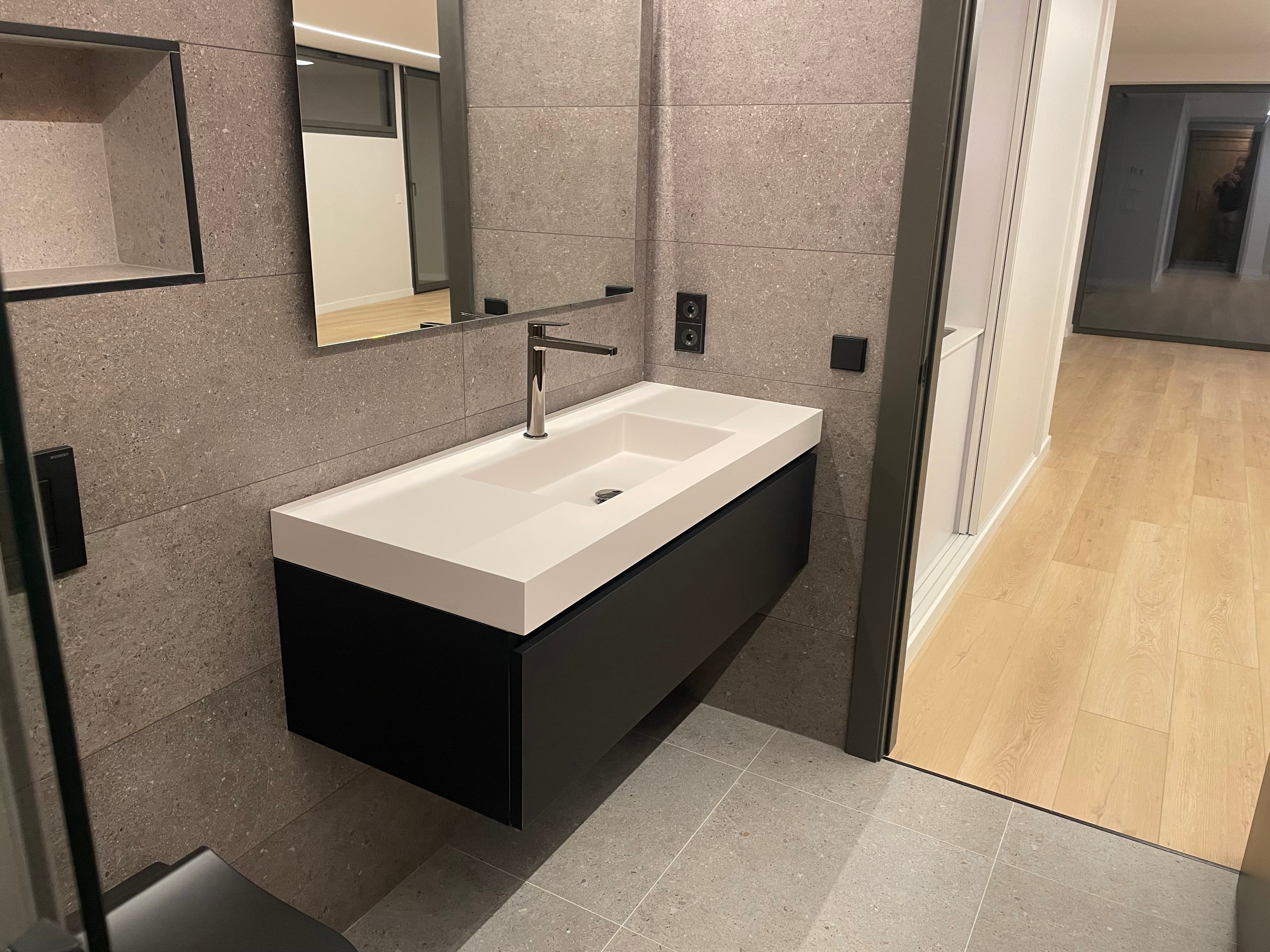Bany modern amb parets i sòl de rajoles grises, lavabo blanc amb moble negre i mirall rectangular.