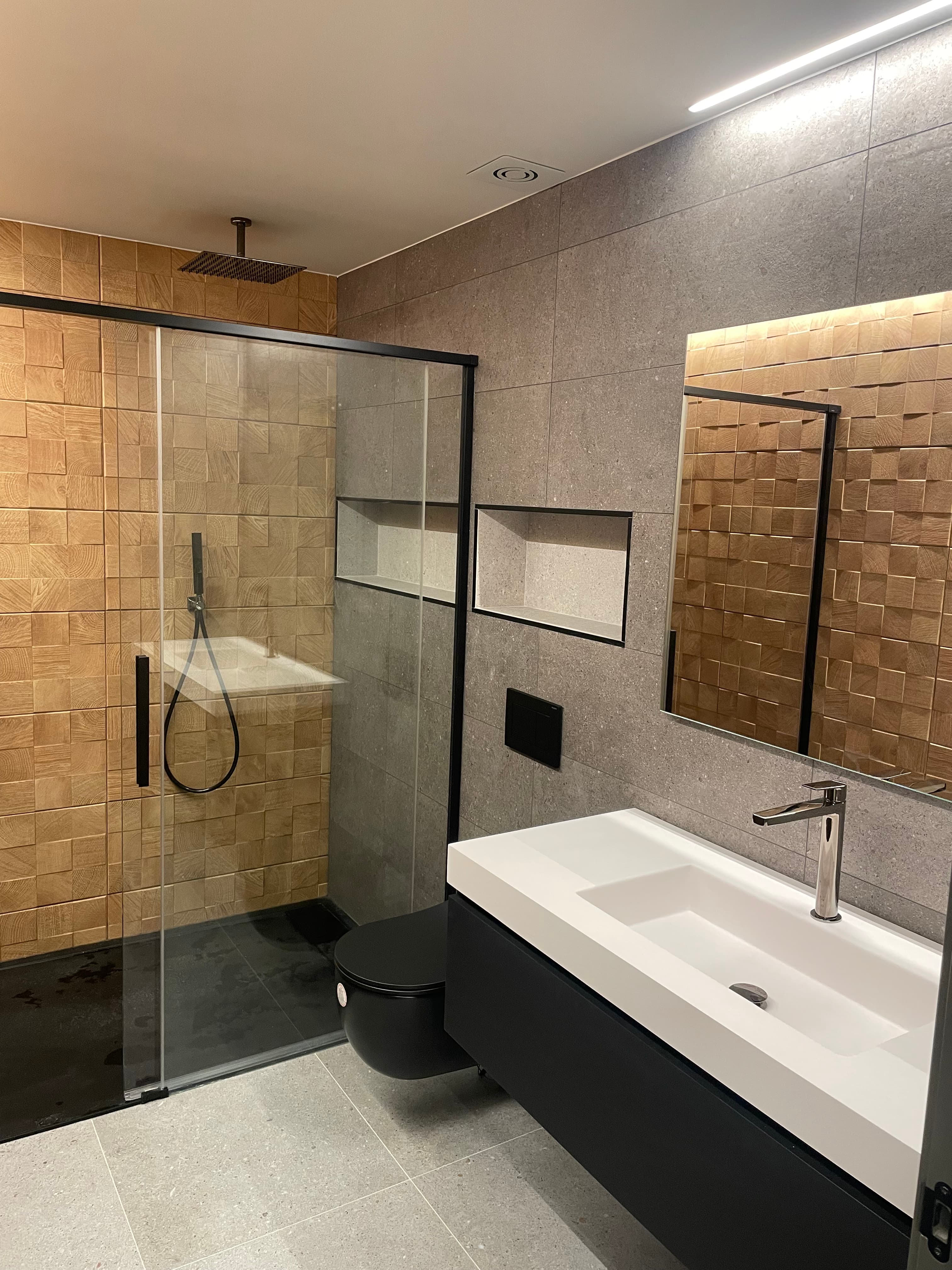 Bany modern amb dutxa de paret de fusta, vàter negre suspès i lavabo blanc amb aixeta metàl·lica i mirall.
