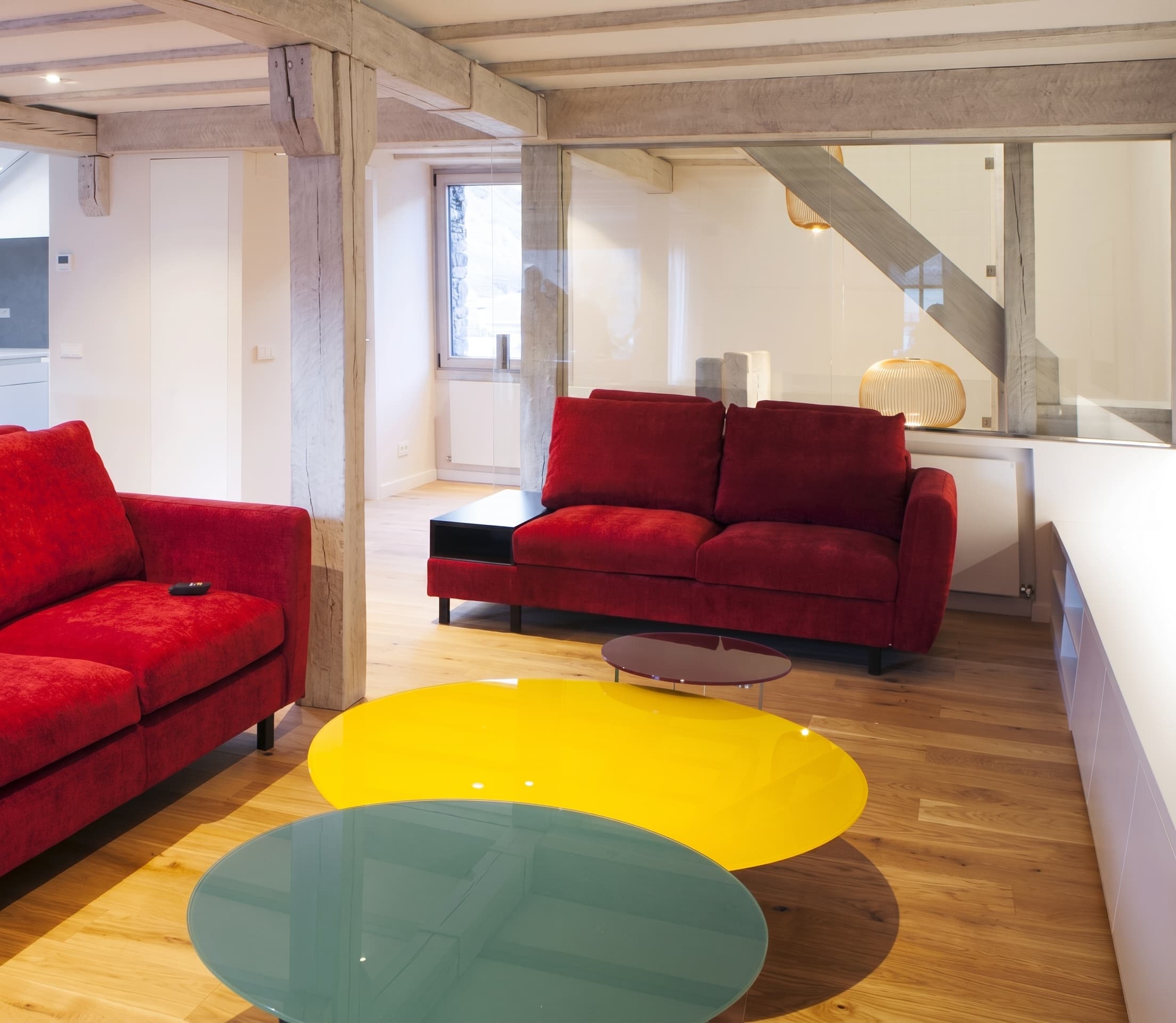 Sala d'estar amb dos sofàs vermells i taules de centre ovalades de colors groc, blau i vermell fosc sobre terra de fusta clara.