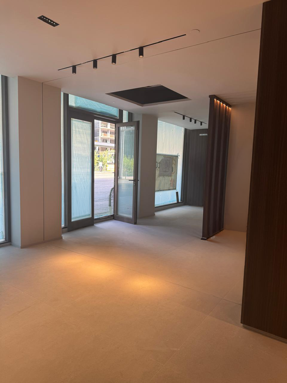 Interior d'una sala buida amb finestrals grans i porta d'entrada oberta, il·luminació de sostre moderna i parets clares amb un racó fosc amb llums puntuals.