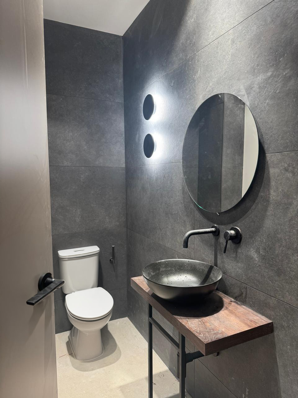 Bany modern petit amb parets fosques, lavabo rodó negre sobre taulell de fusta i vàter blanc.
