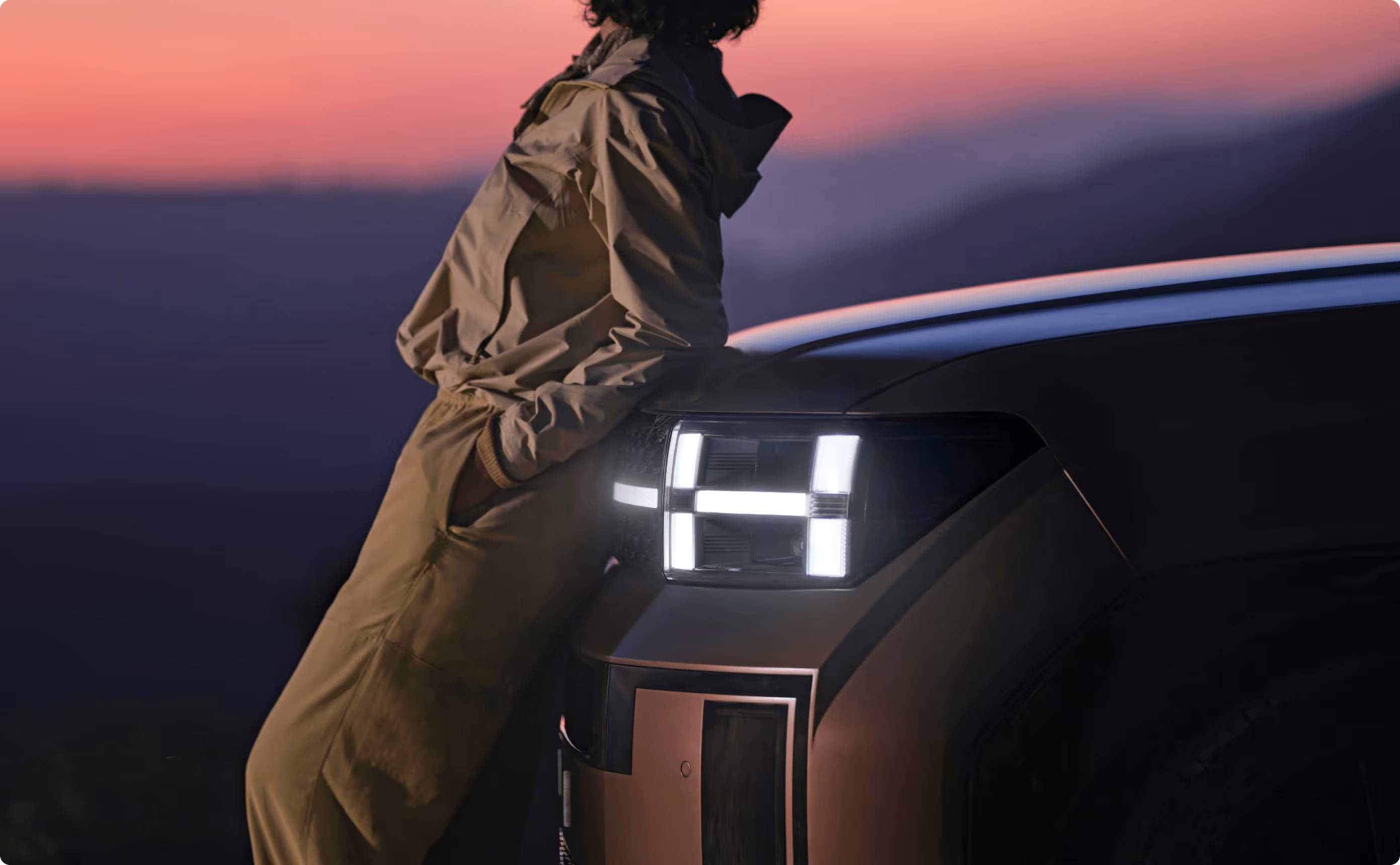 Person in a tan jacket leaning against the front of a vehicle with illuminated modern headlights at sunset.
