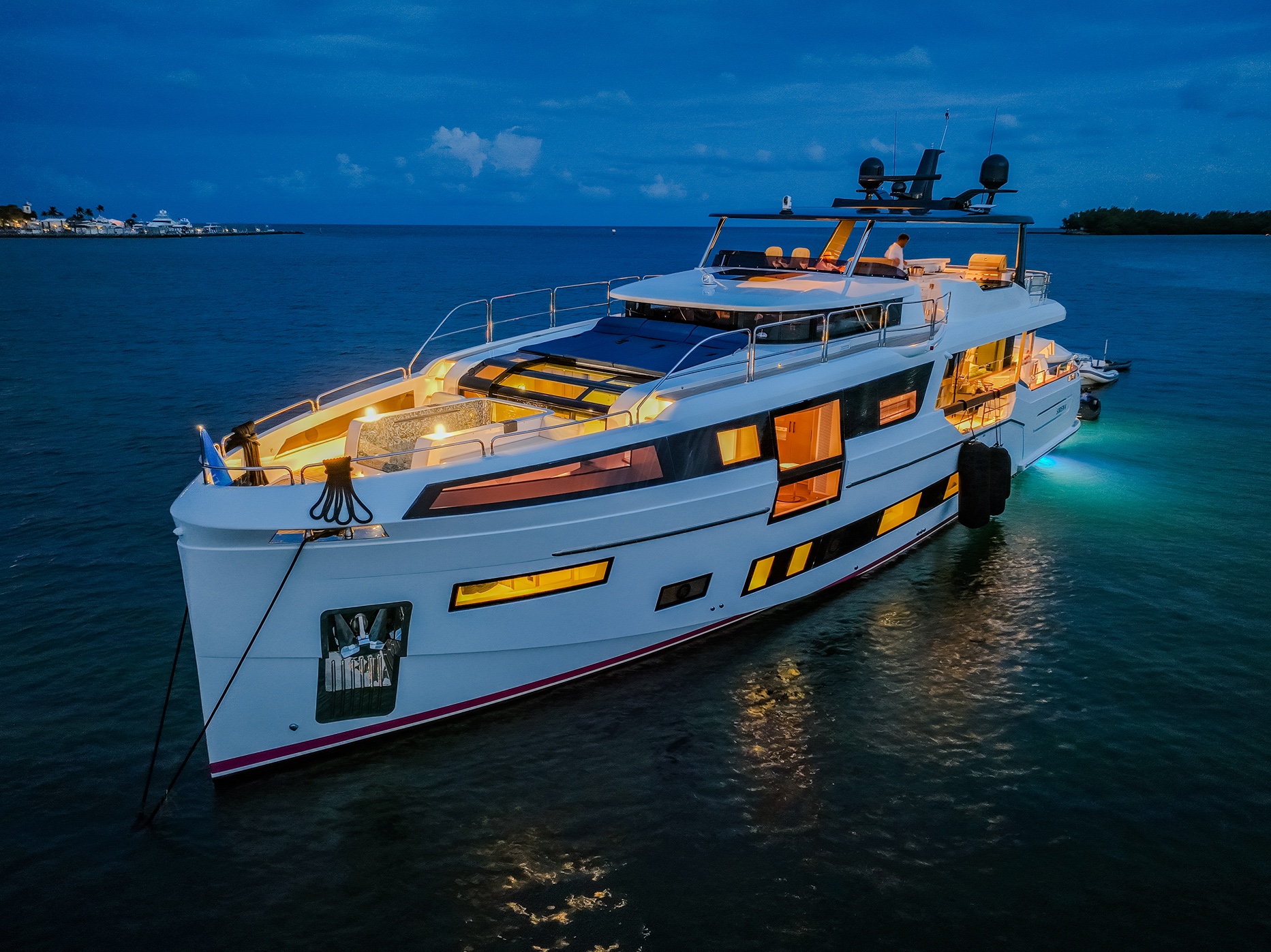 Luxurious white yacht illuminated at dusk floating on calm ocean water with a person on deck.