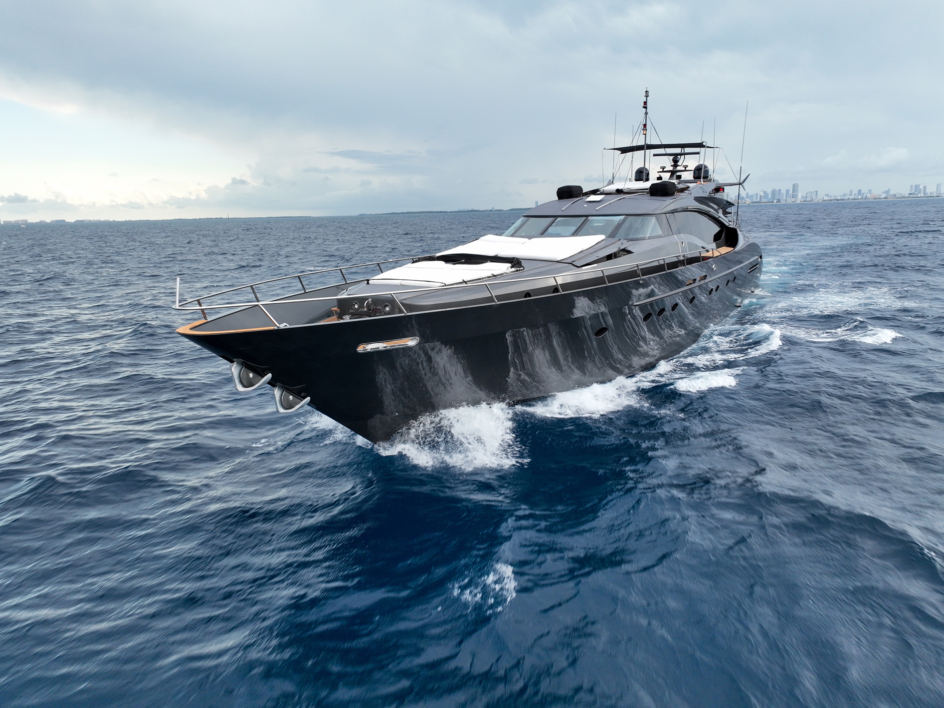 Sleek black luxury yacht cruising on calm blue ocean waters with a distant city skyline under a cloudy sky.
