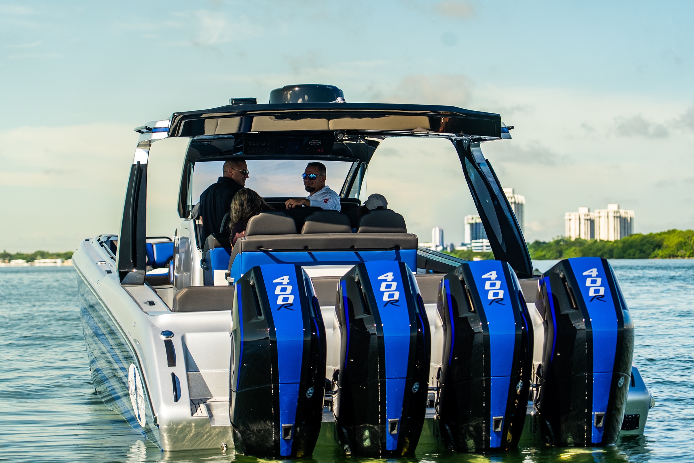 Rear view of a silver powerboat with four blue 400R outboard engines and four people seated inside, near a city shoreline.