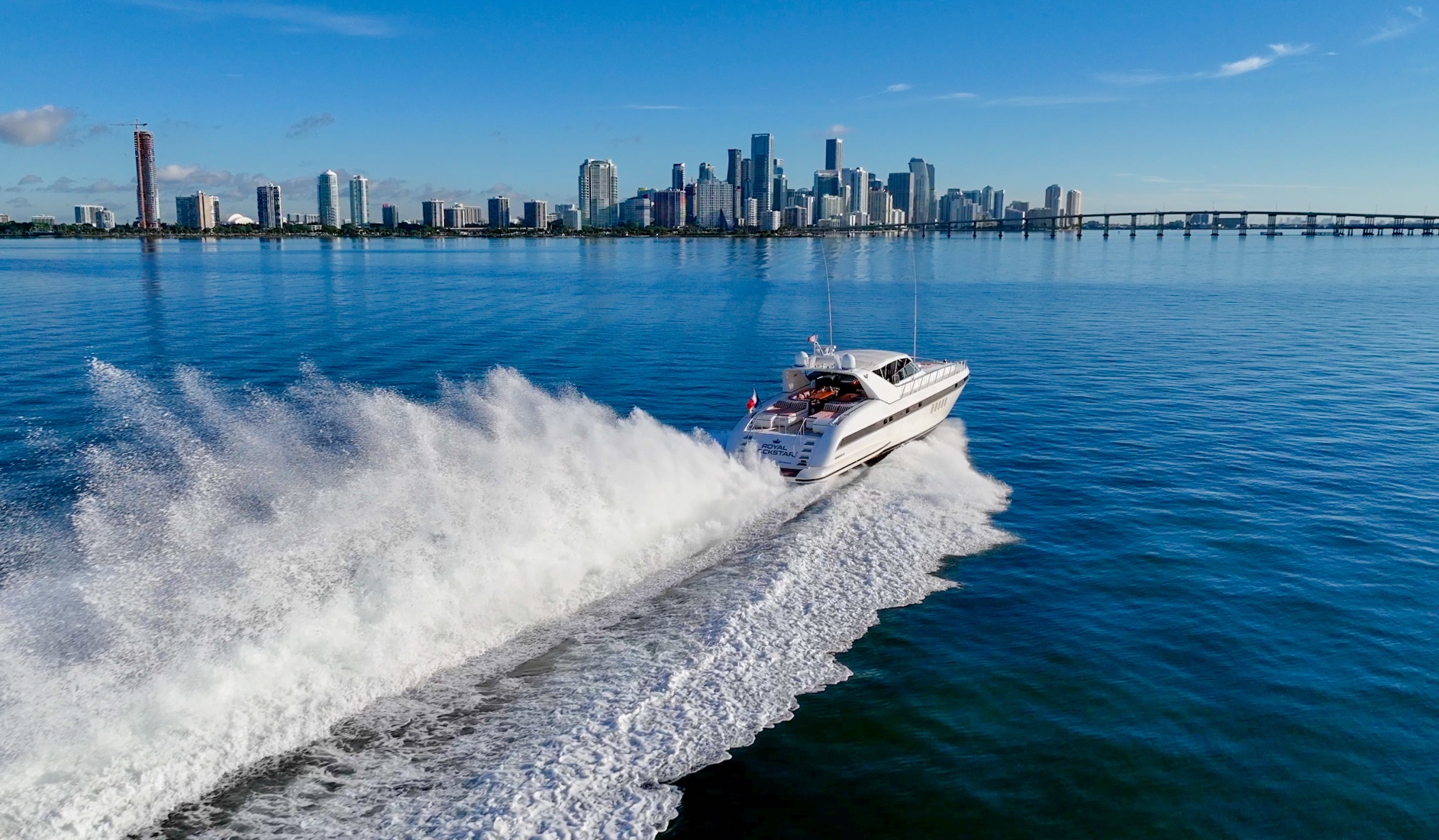 White luxury motorboat speeding across calm blue water with a city skyline and bridge in the background under a clear sky.