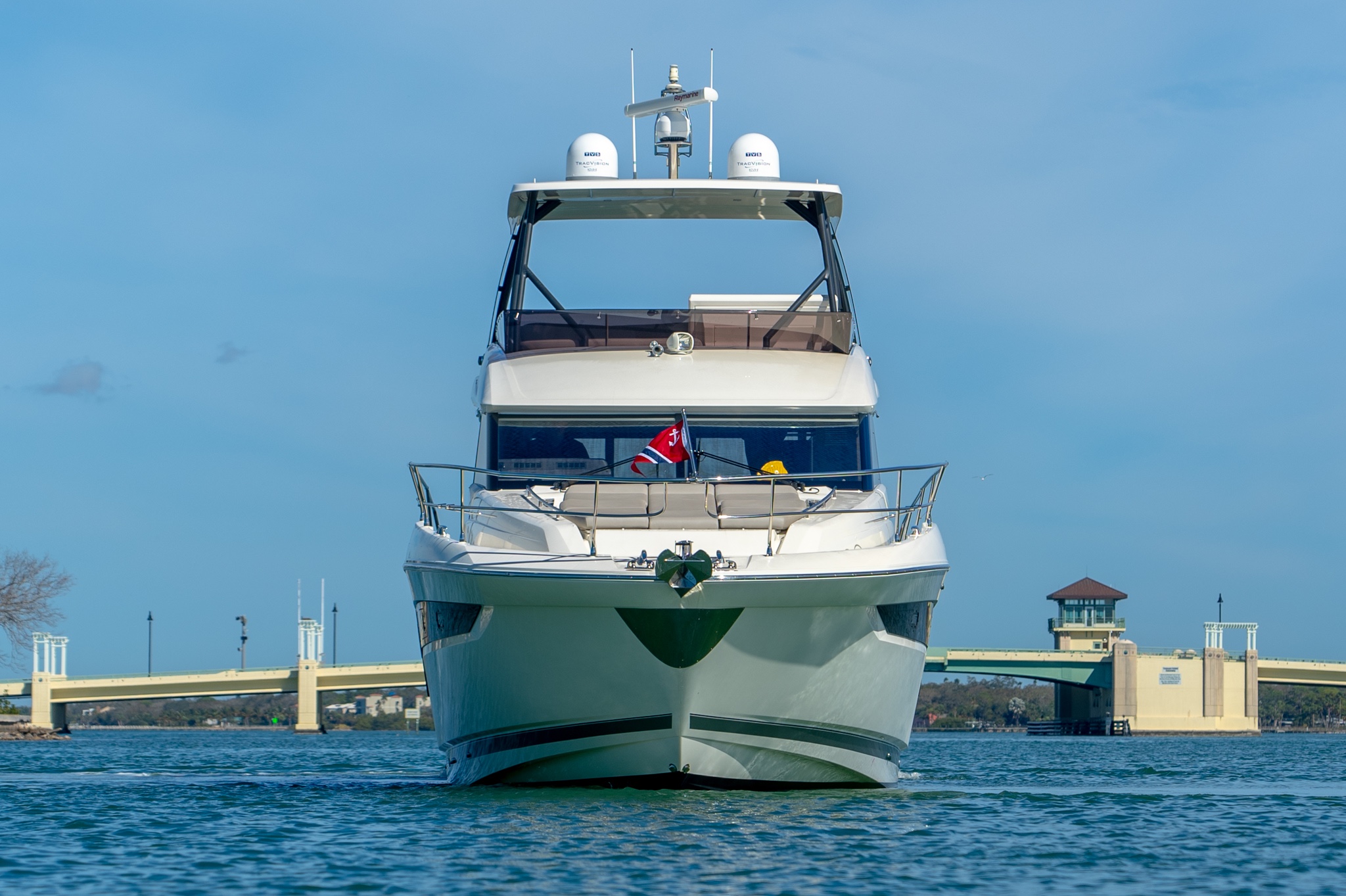 Front view of a white luxury yacht on calm water with a bridge and clear blue sky in the background.