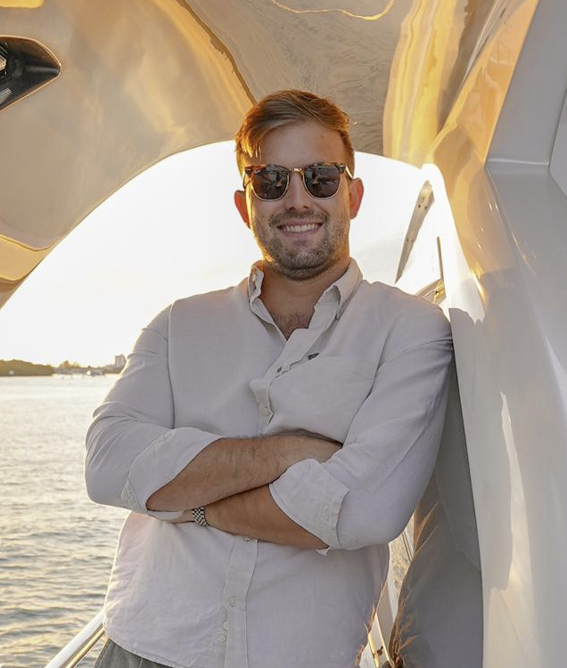 Man wearing sunglasses and a light-colored button-up shirt smiling with arms crossed on a boat, with water and sunset in the background.