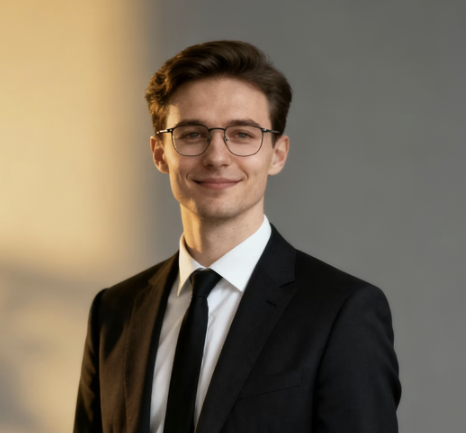 Young man with glasses wearing a black suit and tie, smiling softly against a neutral gray and warm yellow background.