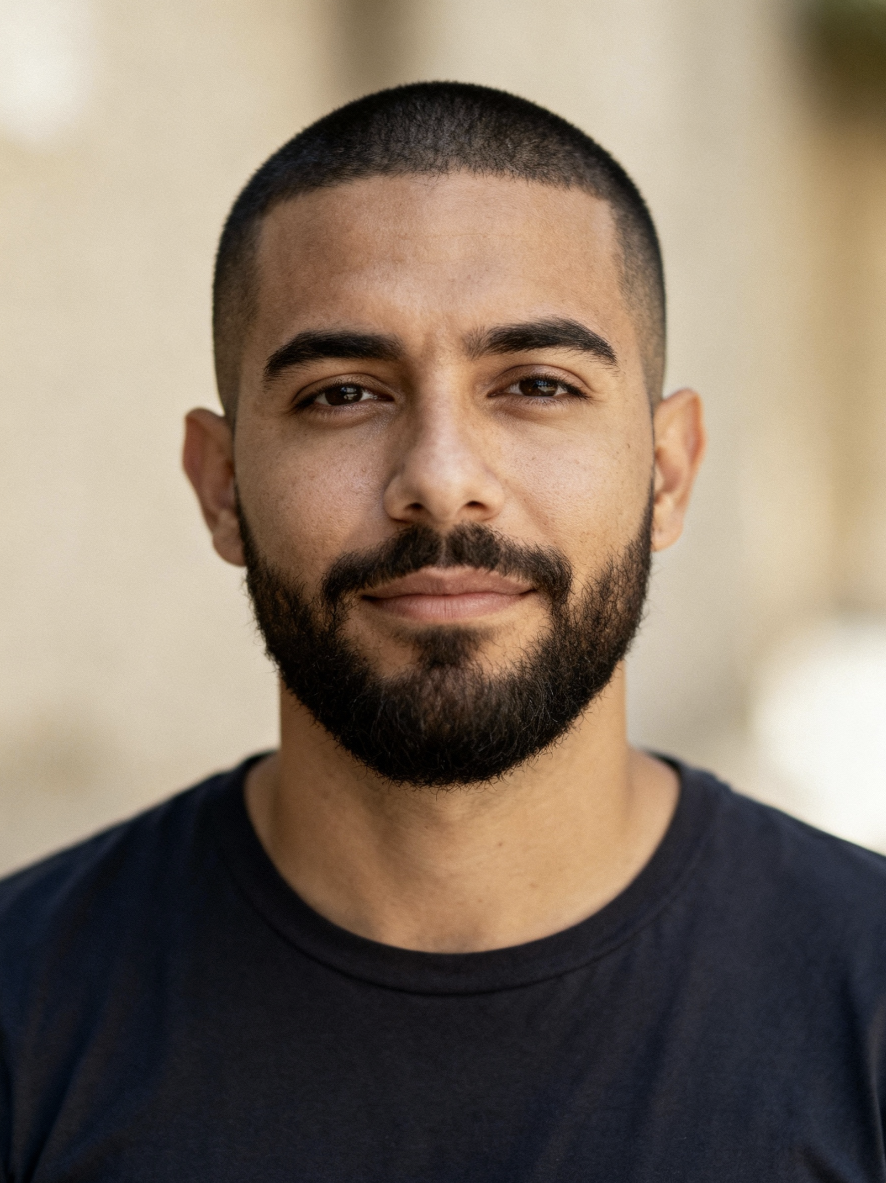 Portrait of a young man with a closely trimmed beard and short hair wearing a dark shirt, looking at the camera with a neutral expression.