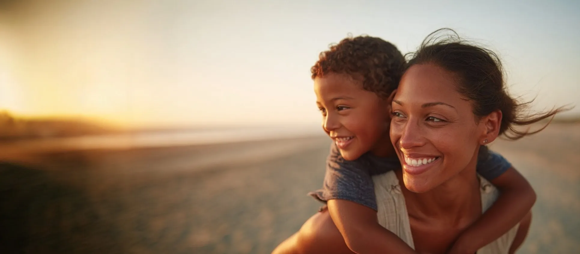Woman carrying child on beach at sunset, both smiling warmly