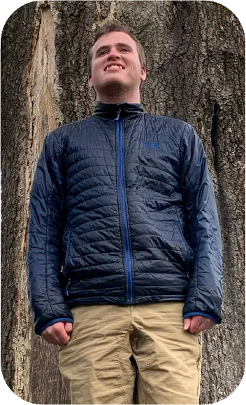 Smiling person in blue jacket standing against large tree trunk outdoors