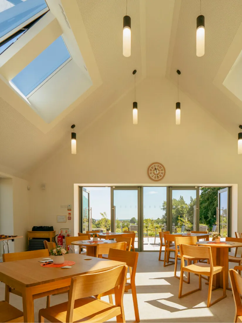 Bright room with wooden tables and chairs, pendant lights, skylights, and glass doors opening to a garden.