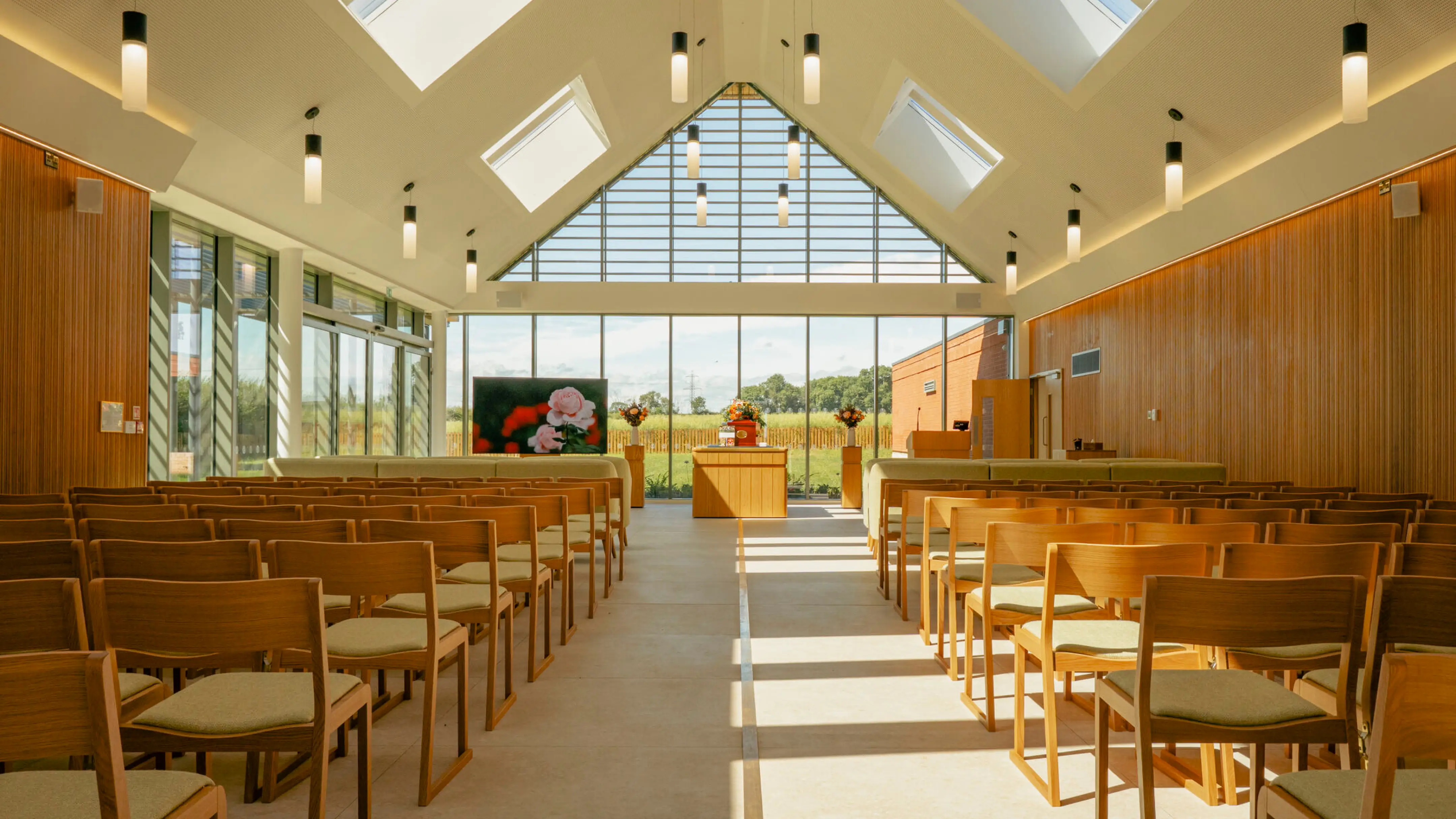 Bright chapel interior with wooden chairs, large glass windows, and a floral display at the front.