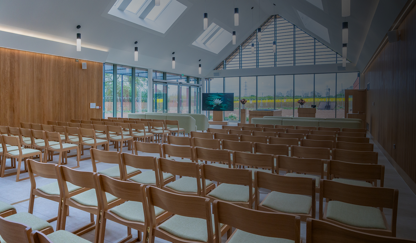 Empty modern chapel with wooden chairs, large windows, skylights, and a TV screen at the front.