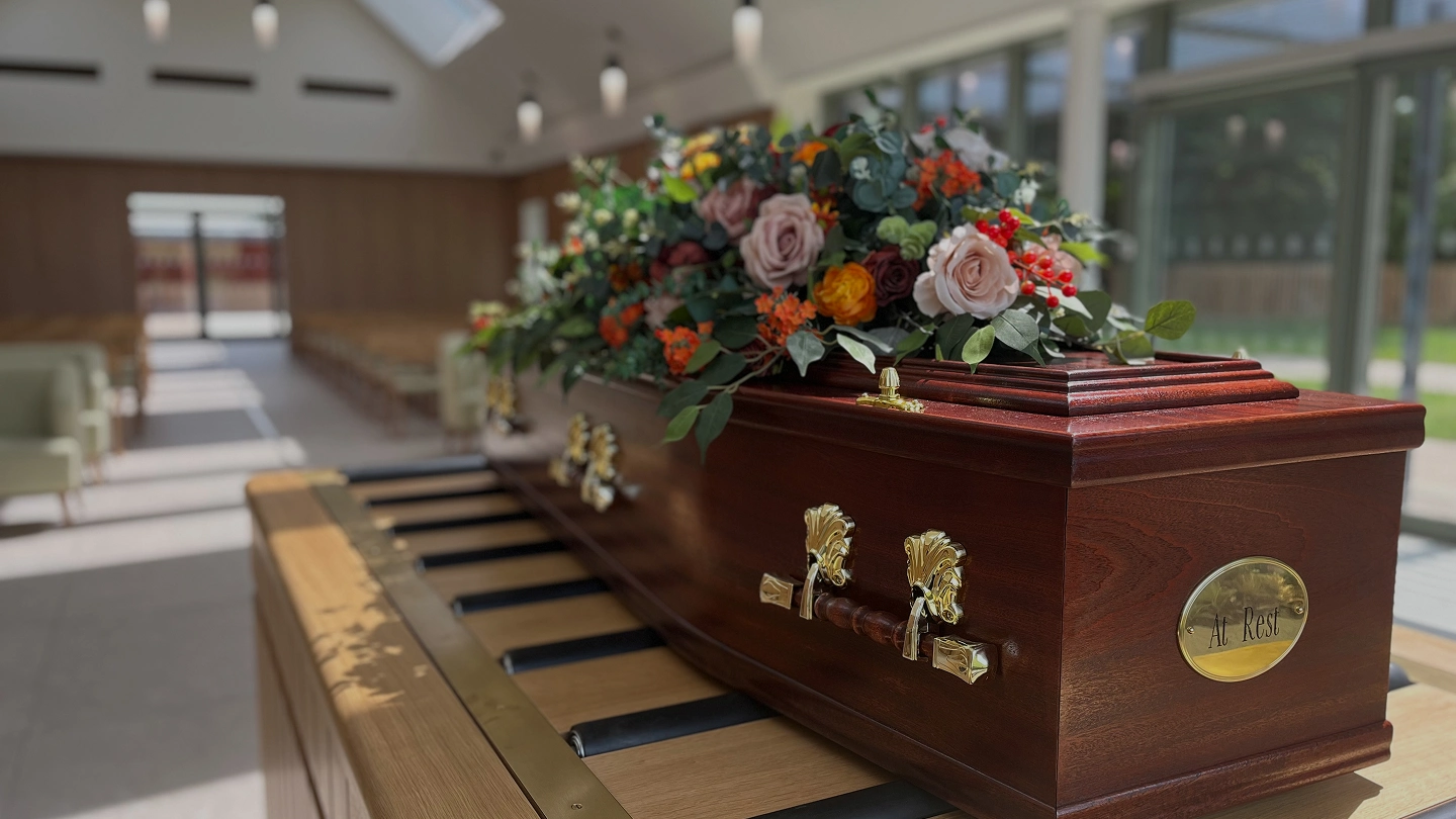 Wooden casket with flower arrangement atop resting on piano keys in a bright room.