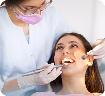 Dentist wearing pink mask and glasses examining smiling woman’s teeth with dental tools.