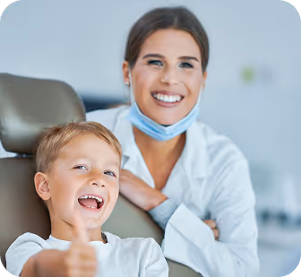 Smiling child giving a thumbs up while sitting in a dental chair with a female dentist behind him.