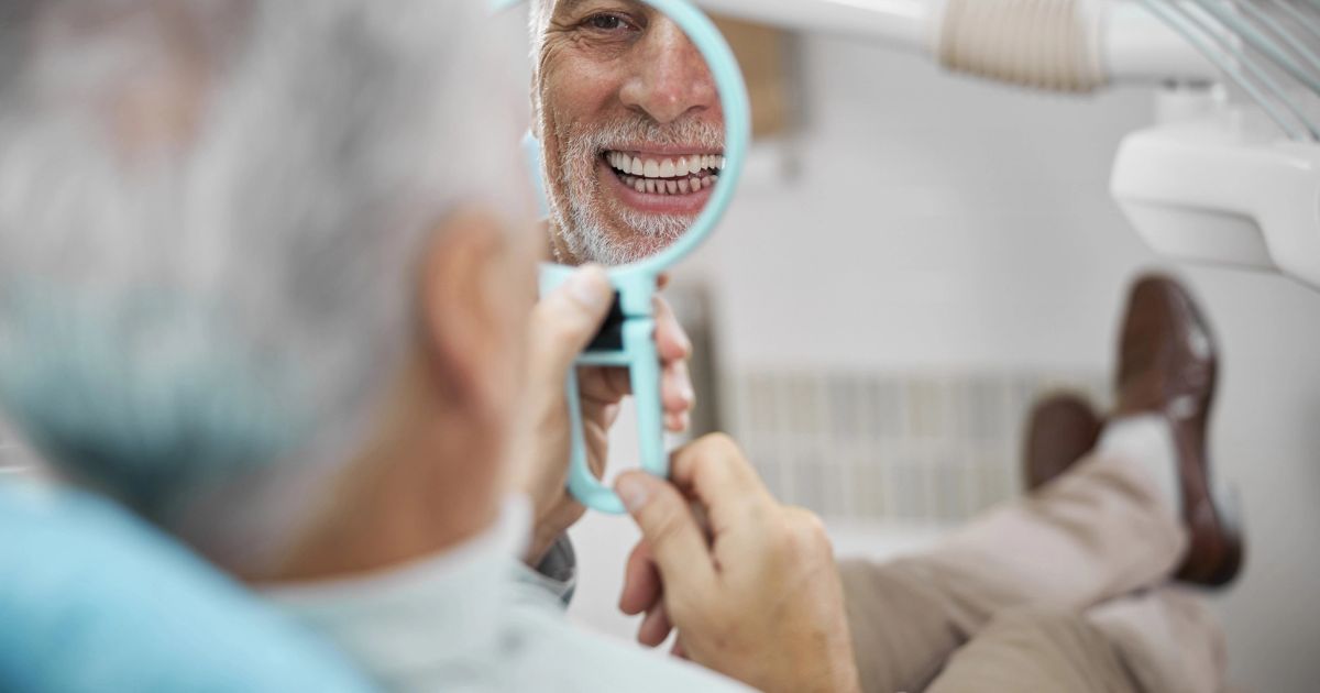 Smiling elderly man with gray hair checking his teeth in a handheld dental mirror while sitting in a dentist's chair.