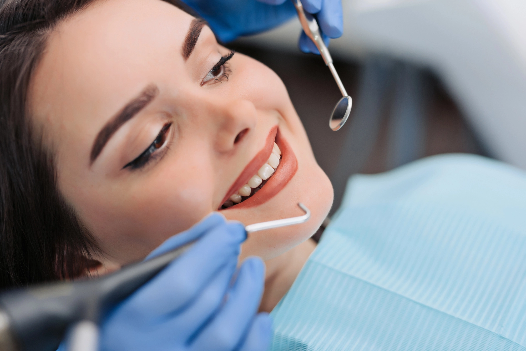 Smiling woman in dental chair during dental examination with tools held by gloved hands.