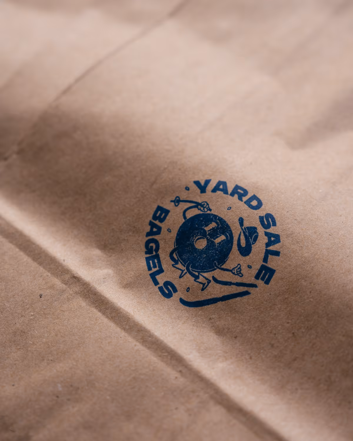 Brown paper bag with a blue logo of a smiling bagel wearing a cowboy hat and crashing on skis, surrounded by the text 'Yard Sale Bagels.'