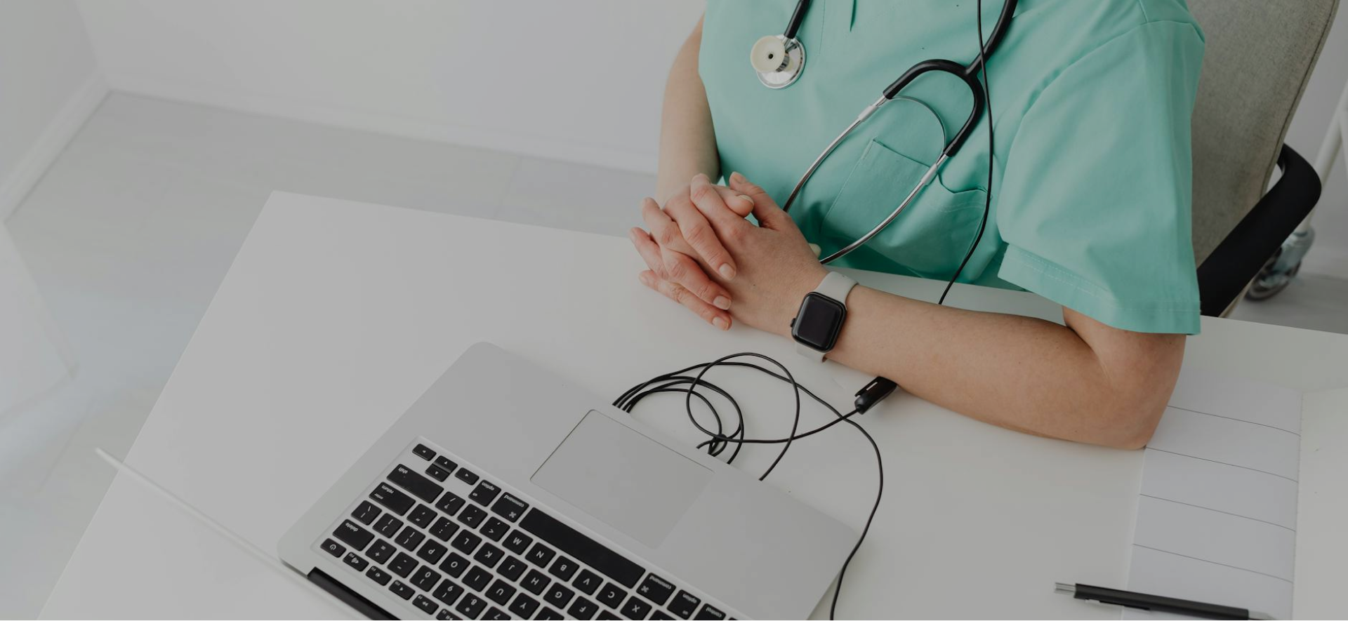 Grayscale image of a nurse sitting at a desk with a laptop and stethoscope.