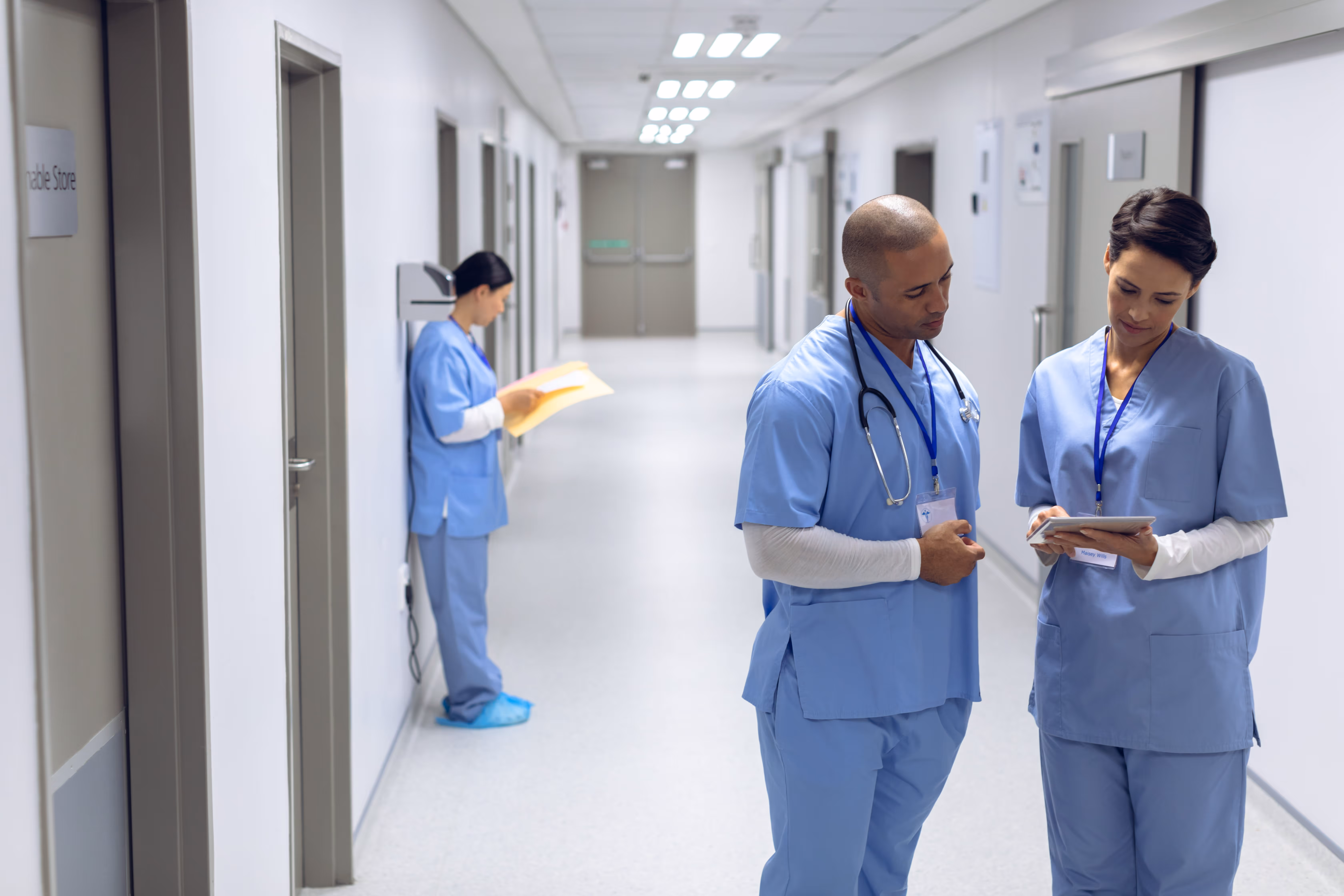 Two healthcare workers in blue scrubs review a tablet while another reads a file in a hospital hallway.