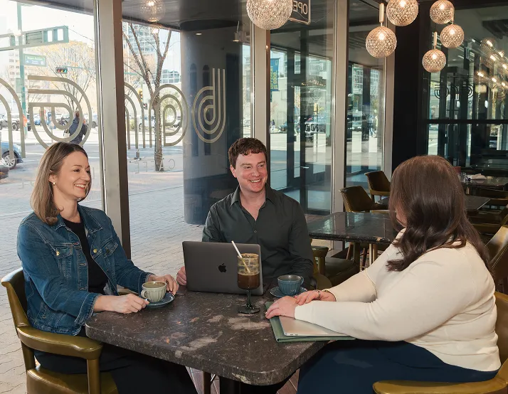 Three people sitting at a café table near window, smiling and conversing with drinks and laptops.