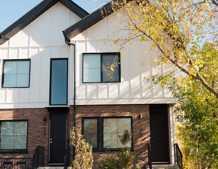 Modern two-story townhouse with white siding, brick lower facade, black-framed windows and doors, and autumn trees around.