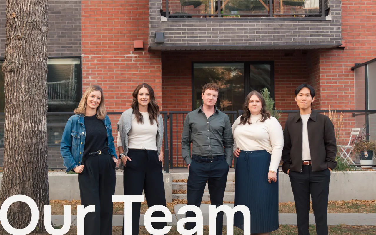 Five professionally dressed people standing outdoors in front of a brick building with a tree on the left, smiling and posing for a group photo.
