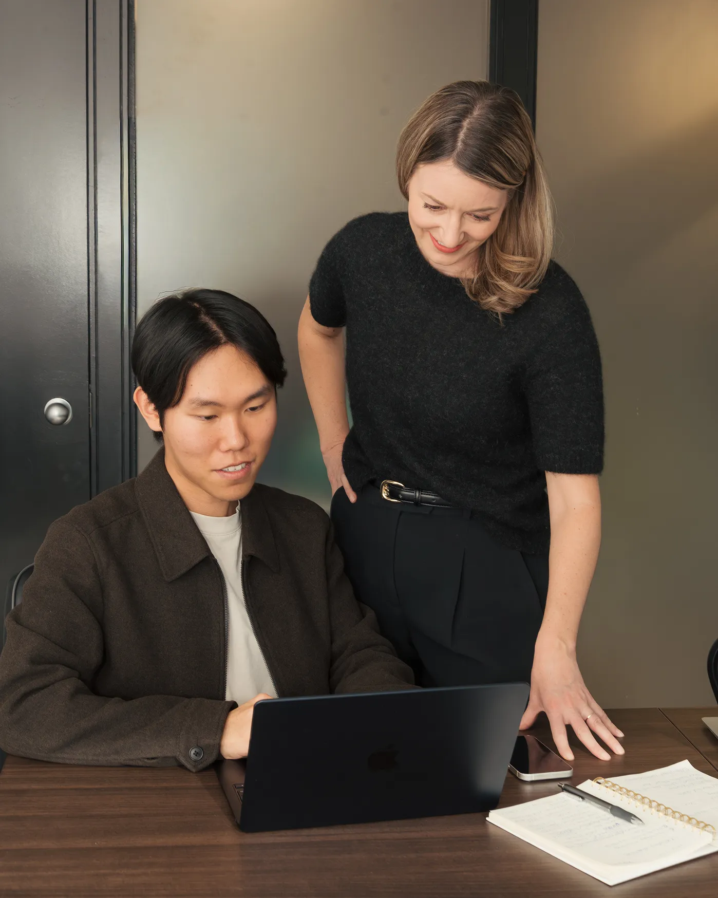 Two colleagues working together at a laptop, with a woman standing and smiling beside a seated man.