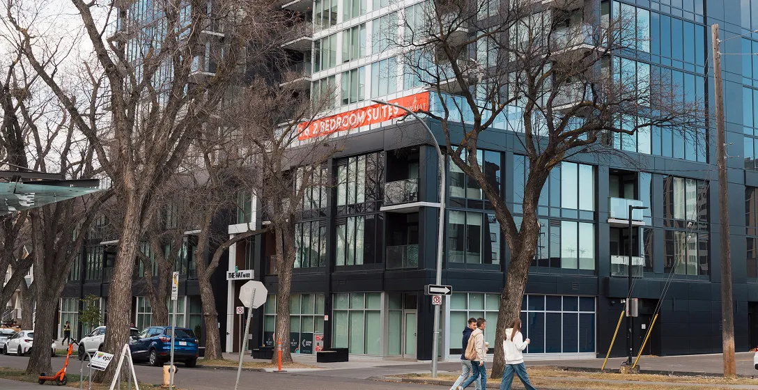 Three people walking on a sidewalk in front of a modern building with large windows and leafless trees along the street.