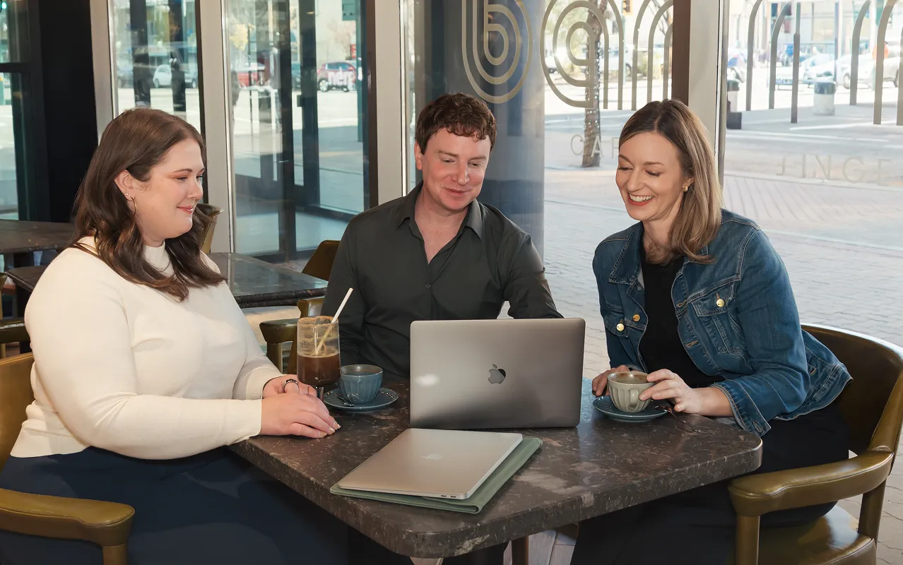 Three people sitting at a cafe table with laptops and drinks, looking at a screen and smiling.