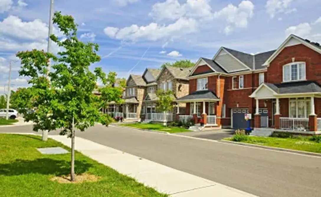 Suburban street with modern single-family houses, green lawns, and a young tree under a partly cloudy sky.