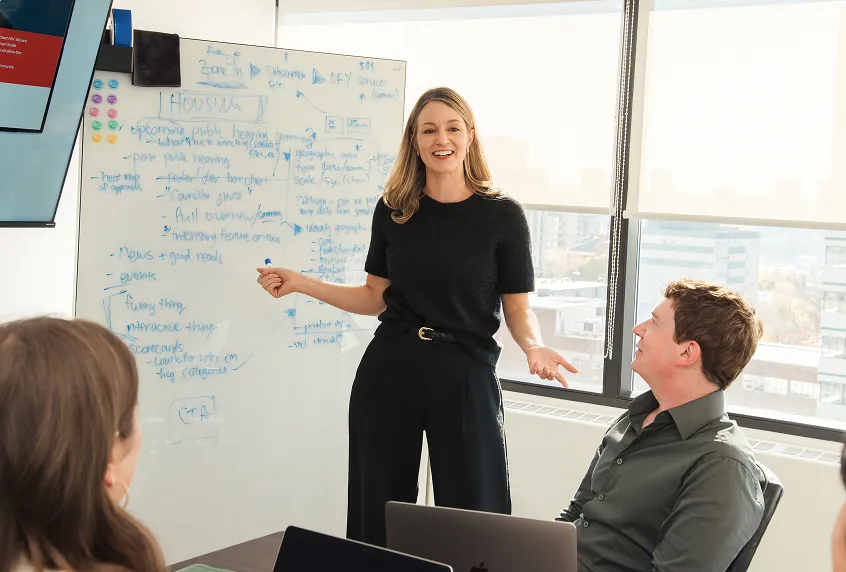 Woman in black shirt standing and presenting in front of a whiteboard to colleagues seated around a table in a bright office.