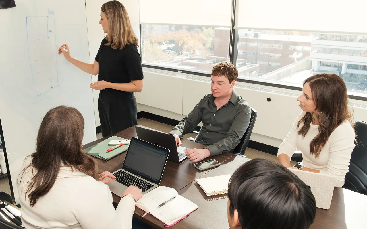 A woman stands writing on a whiteboard while four colleagues sit at a table with laptops and notebooks in a bright office.