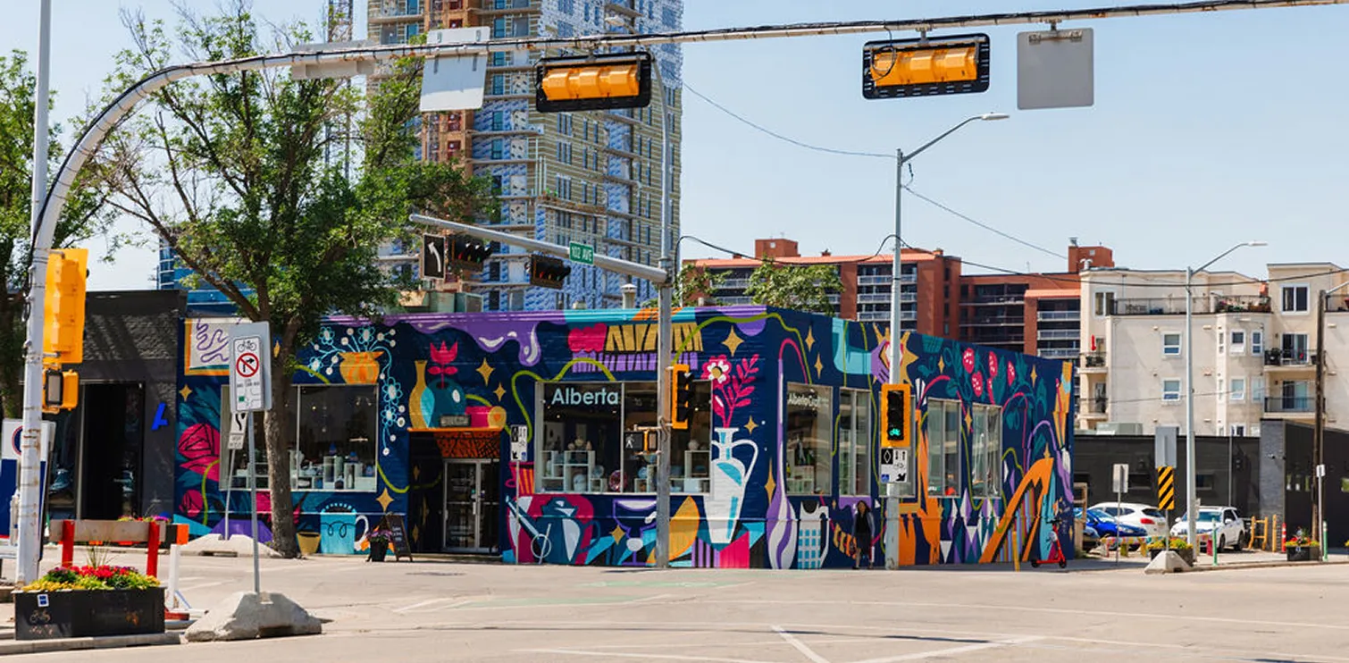 Colorful mural with abstract shapes and plants painted on a corner building named Alberta in a city setting with traffic lights and nearby high-rise buildings.