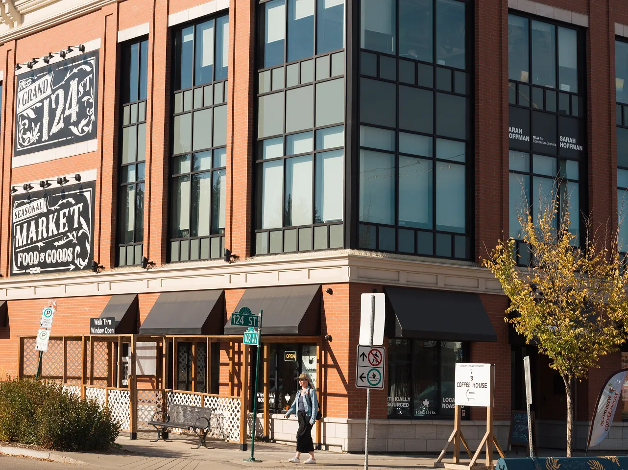 Corner of a brick commercial building with large windows featuring signs for Grand 124 St Seasonal Market Food & Goods and a coffee house, with a woman walking on the sidewalk.