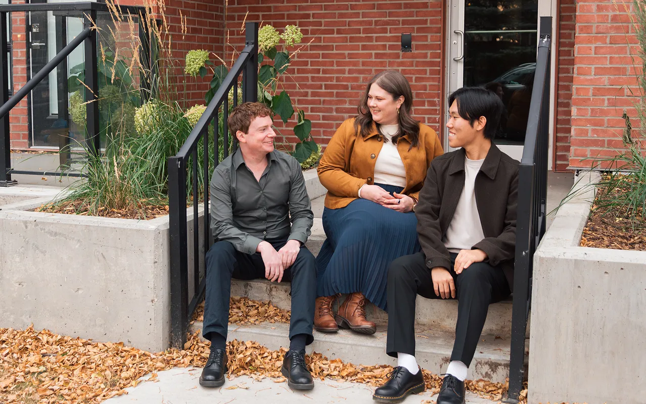 Three people sitting on concrete steps outside a brick building, smiling and engaging in conversation.