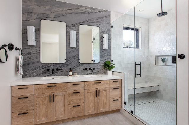 Modern bathroom featuring a double vanity with natural wood cabinets, marble backsplash, dual mirrors, and a glass-enclosed walk-in shower. Designed by Denver Dream Builders.