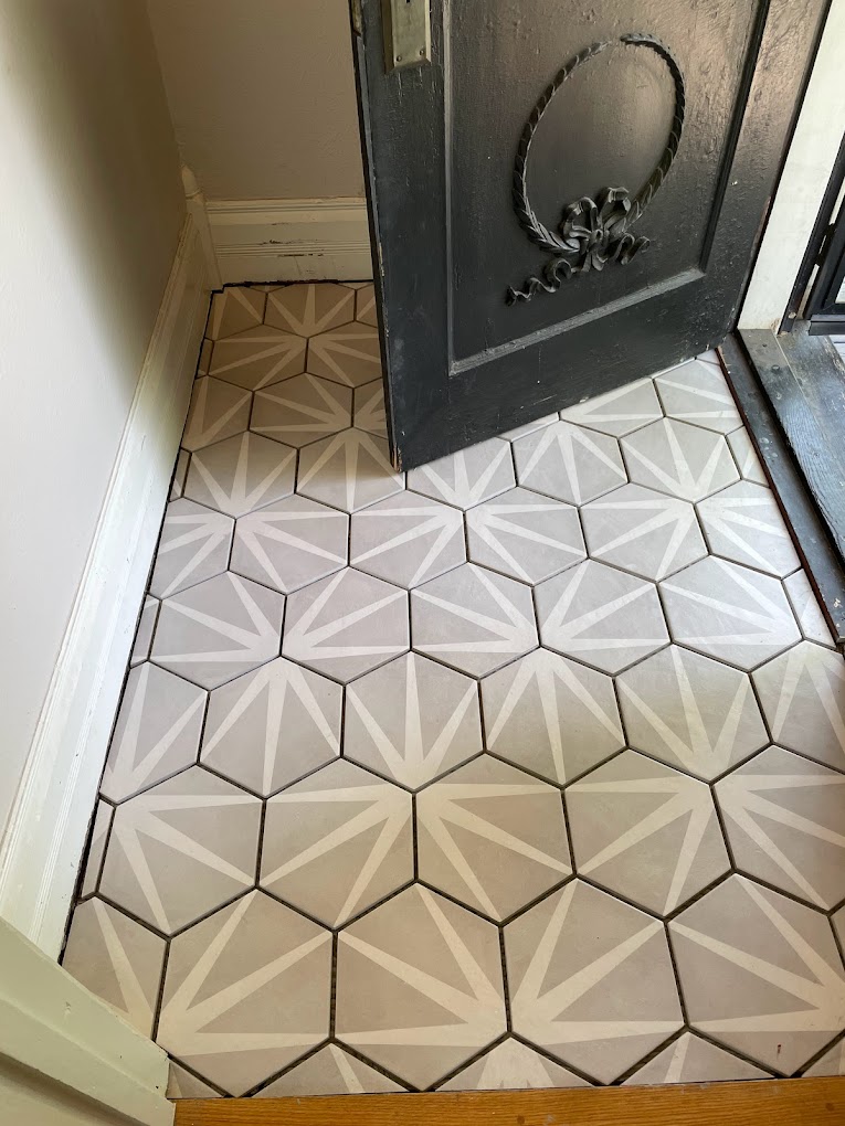 Intricate geometric tile flooring in a neutral palette, leading to a decorative black metal door with ornate detailing.