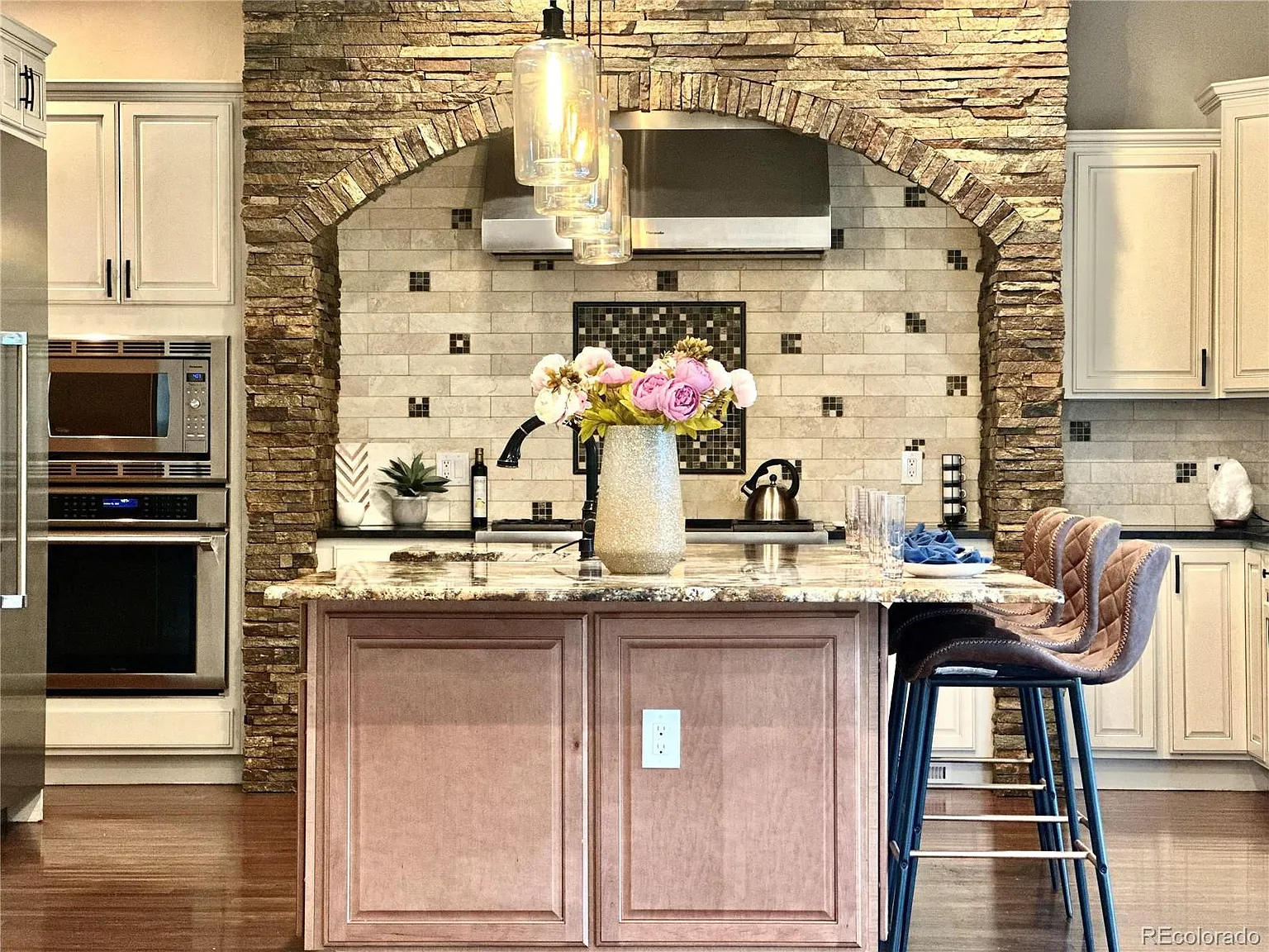 Rustic kitchen design featuring a stone archway over a tiled backsplash with a mosaic accent, stainless-steel range hood, and warm wood cabinetry. A granite island with flowers in a vase and pendant lighting adds charm to the space.