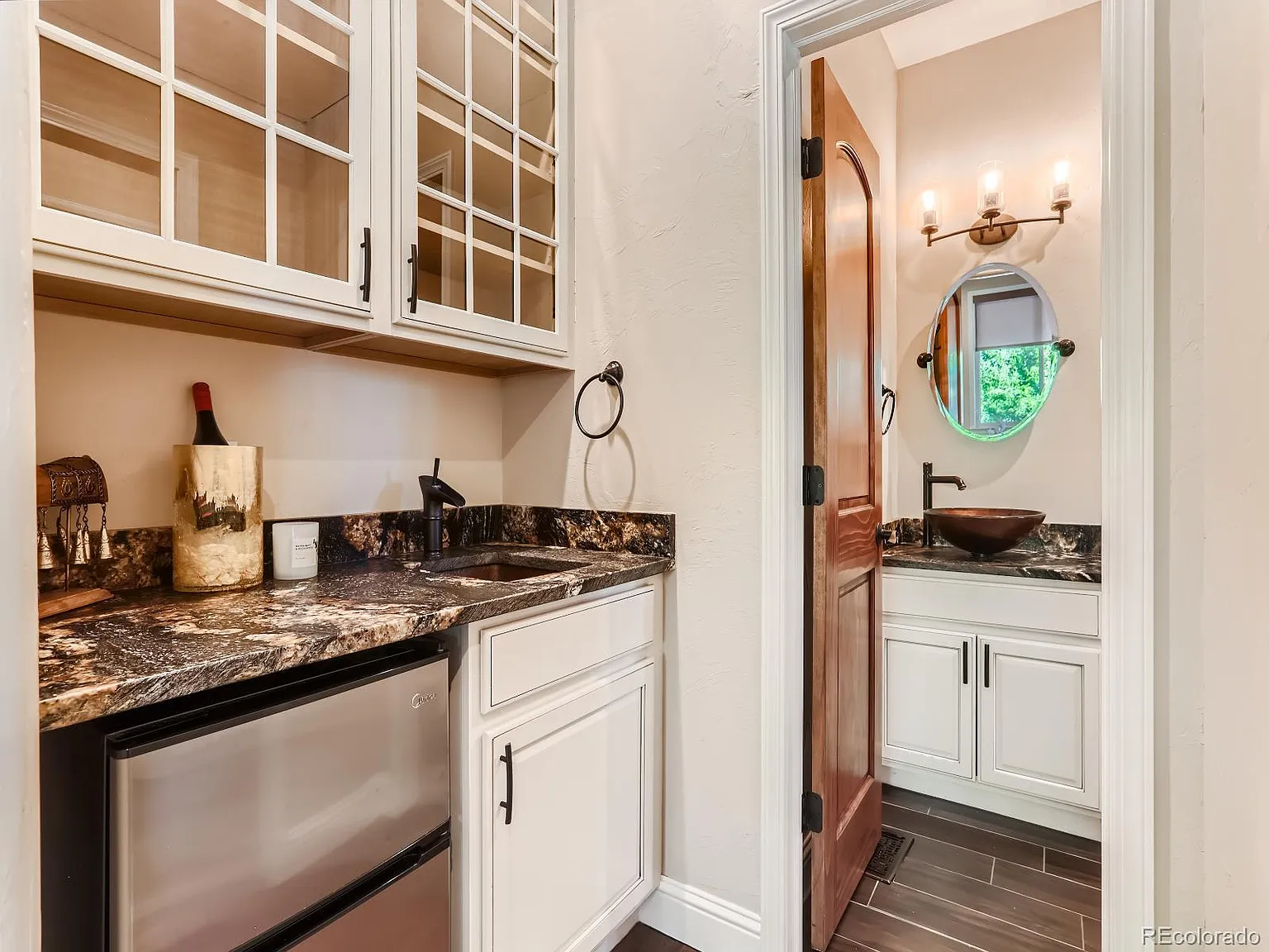Compact bar area with glass-front cabinetry, granite countertops, and a built-in beverage fridge, leading to a small bathroom featuring a vessel sink, a wooden door, and a round mirror with stylish lighting.