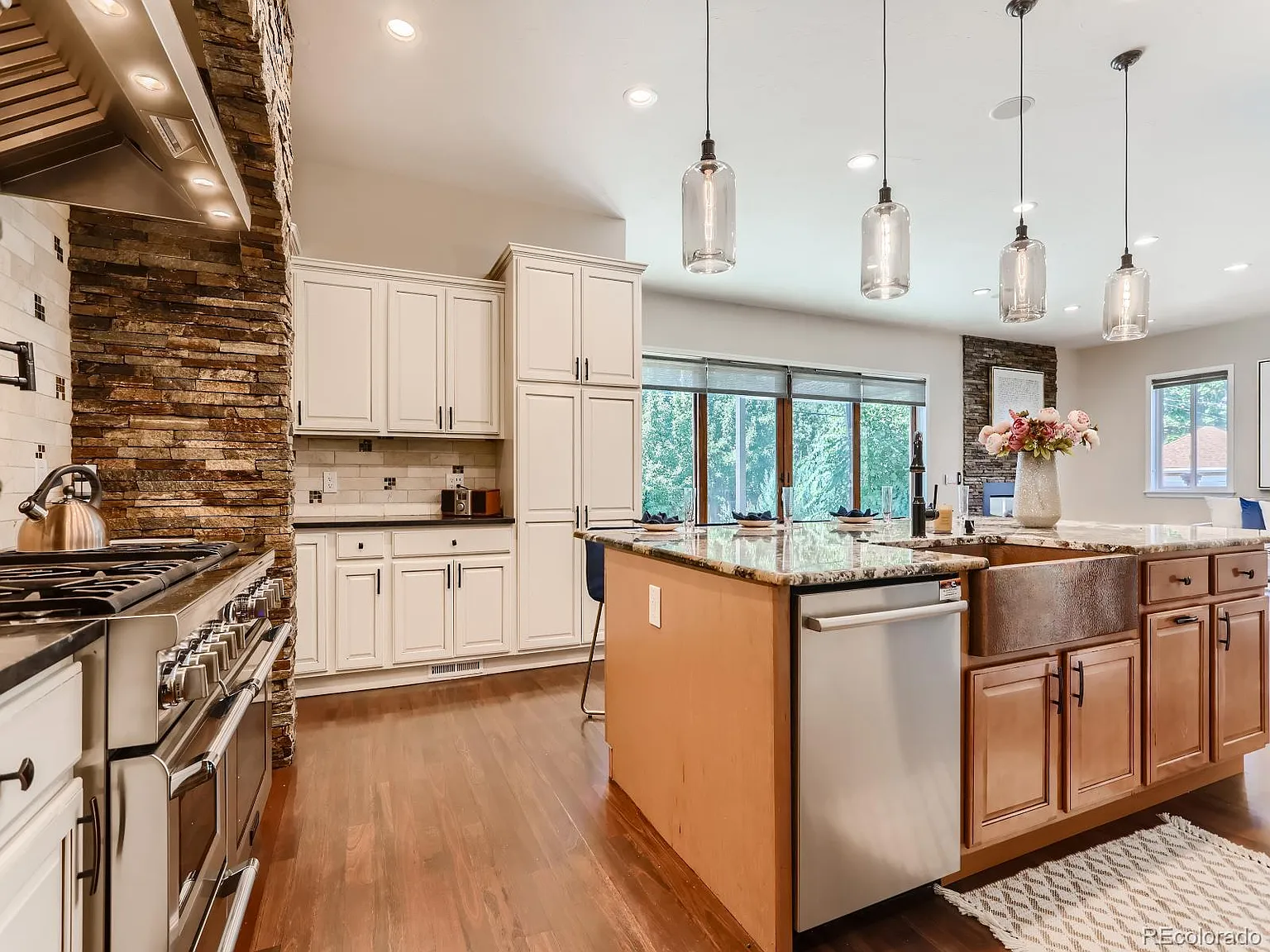 Spacious kitchen featuring a stone backsplash with a large range, white cabinetry, a center island with a wood finish, granite countertops, modern pendant lighting, and hardwood flooring, overlooking a bright dining area with large windows.