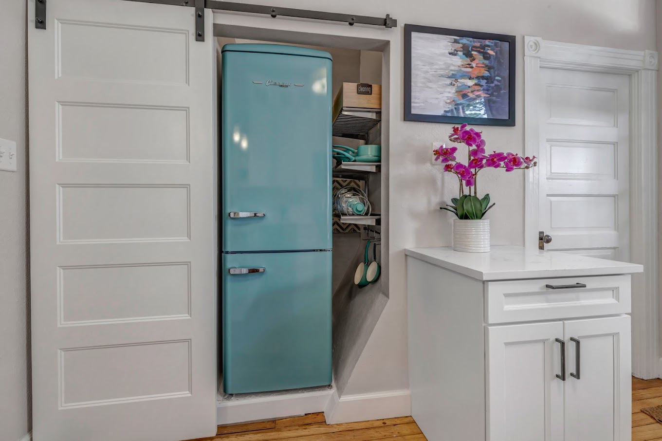 Modern kitchen with a retro-style turquoise refrigerator behind sliding barn doors, a small white island with storage, and a vibrant pink orchid centerpiece.